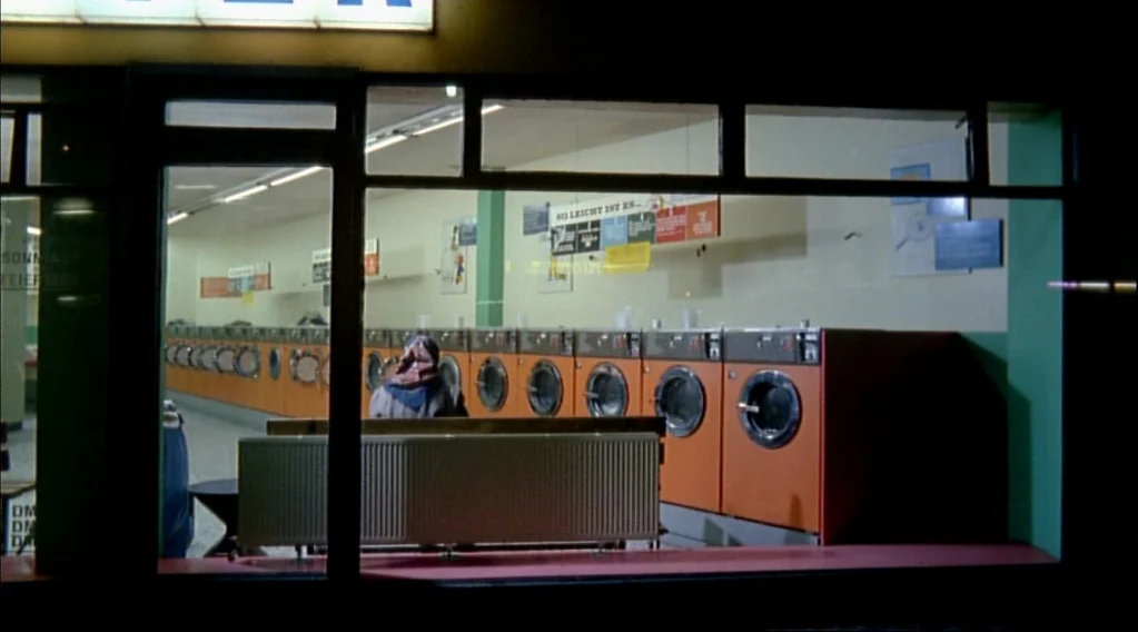Wings Of Desire (1987) movie still: wide — A quiet laundromat at night with a row of orange machines along the wall. A lone person sits on a bench,…