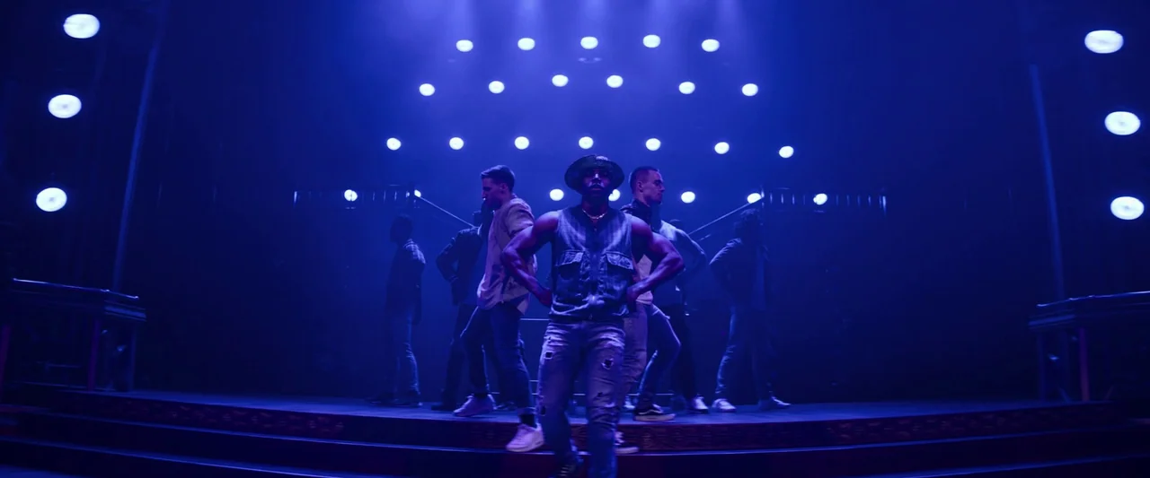 Magic Mike's Last Dance (2023) movie still: fullbody — A group of dancers stands on a darkened stage illuminated by a grid of lights. The performers…