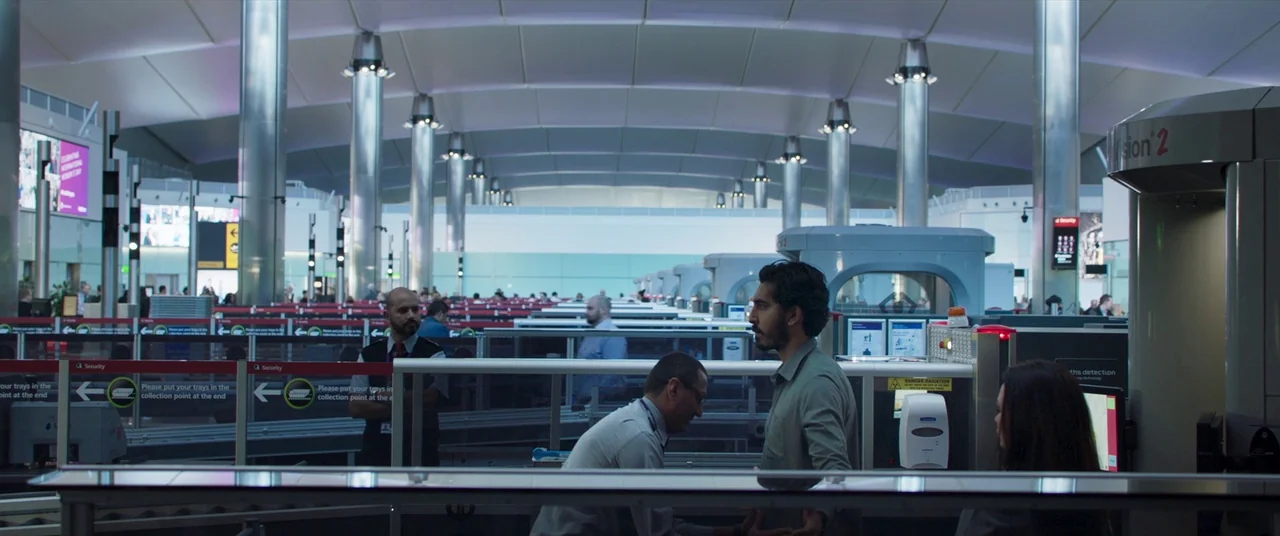The Wedding Guest (2018) movie still: wide — A wide view of a modern airport security screening area with metal scanners. Passengers and security…