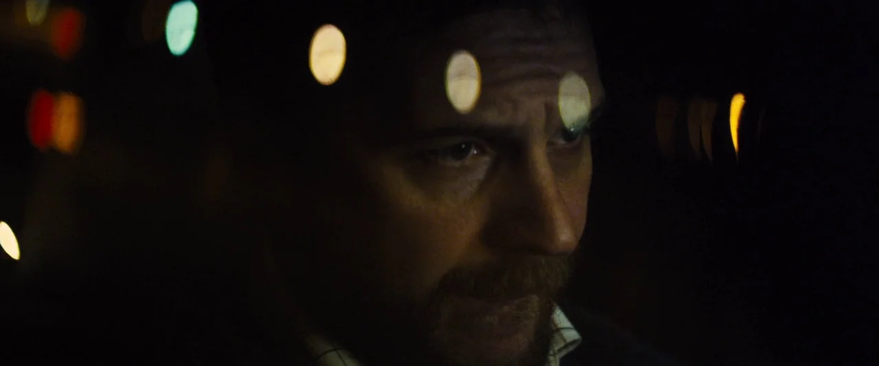 Locke (2013) movie still: closeup — A man sits inside a dark vehicle at night. Blurred bokeh lights are scattered across the dark foreground and…