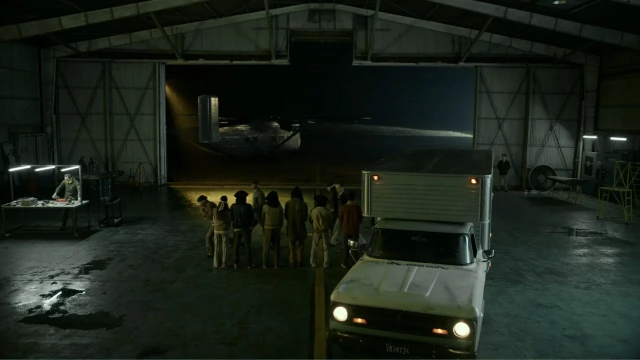 Transfers (2024) movie still: wide — A dark, expansive aircraft hangar with a large open bay door revealing a parked plane. A small group of people…