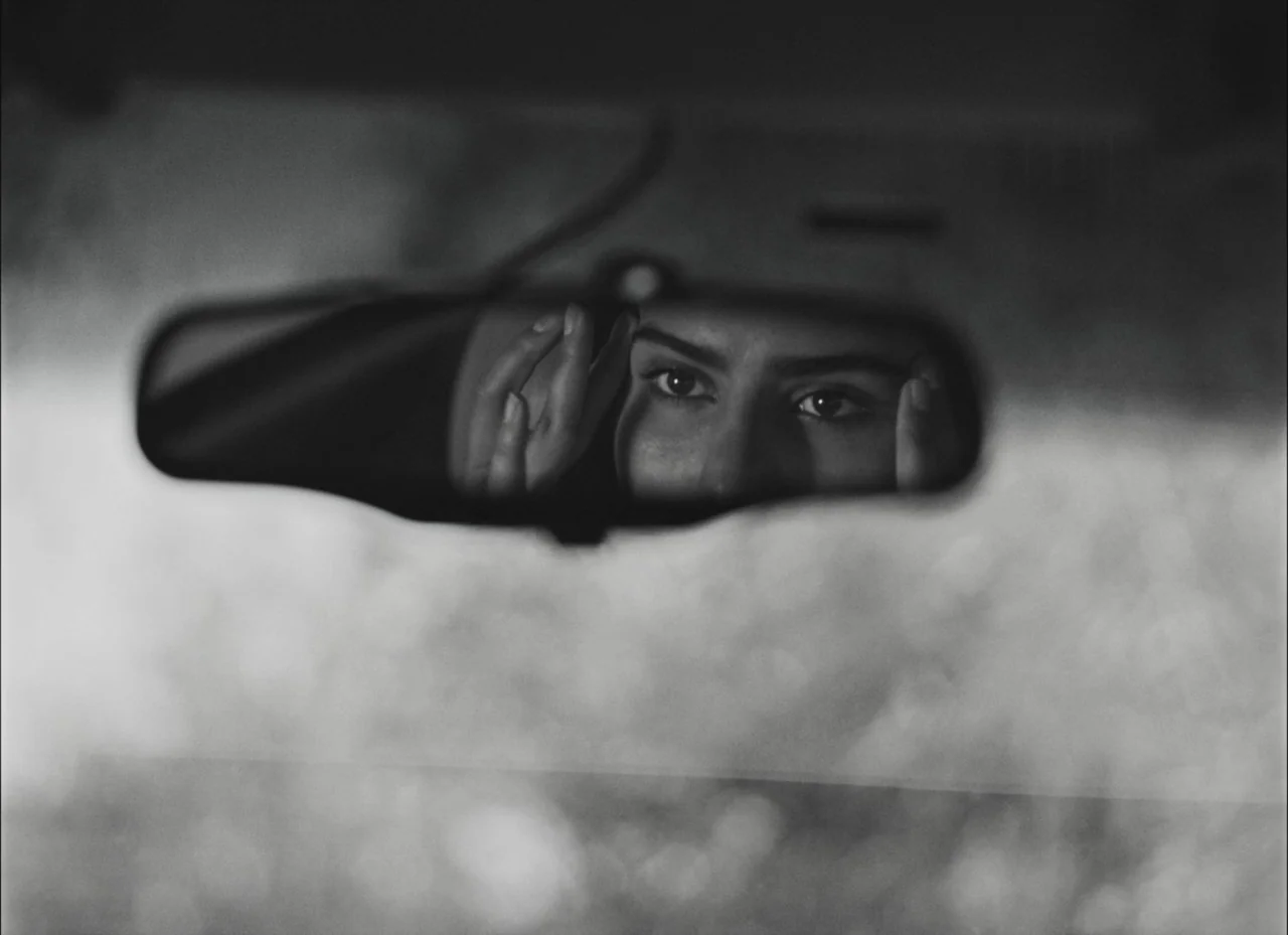 Fremont (2023) movie still: closeup — The interior of a car at night seen through the reflection in the rearview mirror. A young woman looks at her…