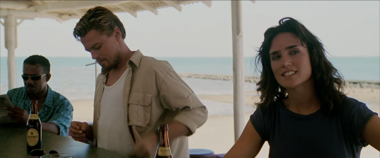 Blood Diamond (2006) movie still: medium — An open-air beach bar with white support pillars stands against a bright sea background. A young man, a…