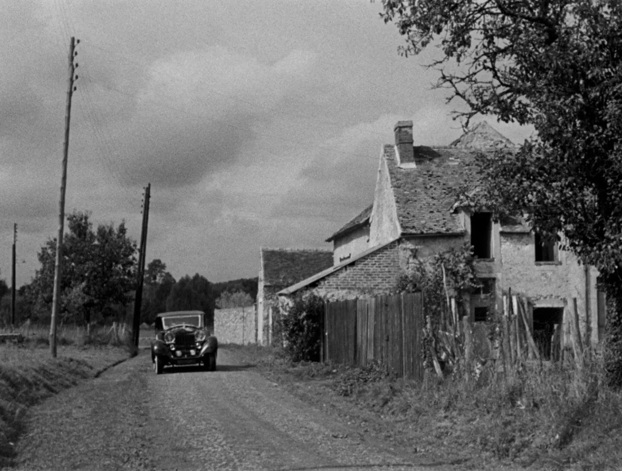Orpheus (1950) movie still: wide — A quiet rural road passes by an aging stone house with a wooden fence. A vintage automobile drives down the…