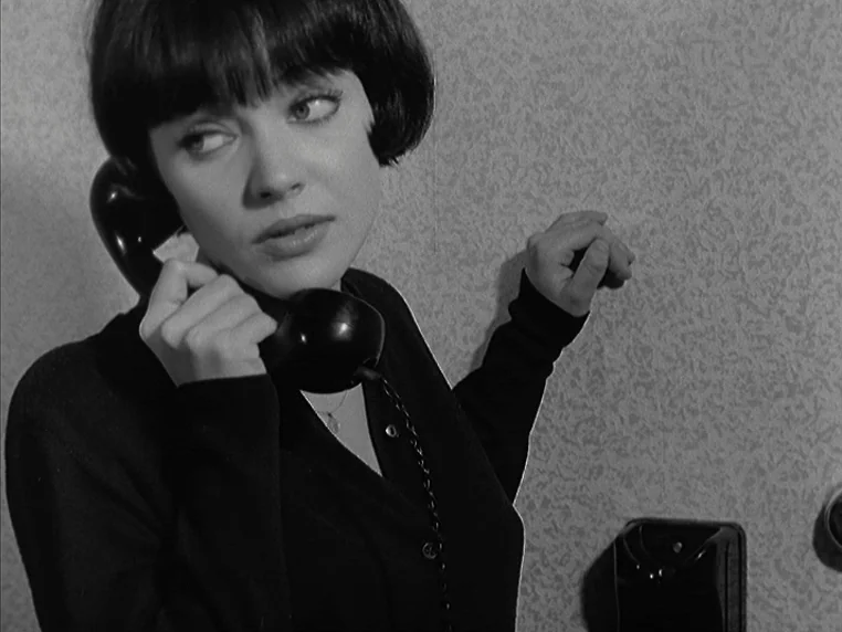 Vivre Sa Vie (1962) movie still: closeup — A young woman stands against a textured wall inside a simple indoor space. She holds a black vintage…