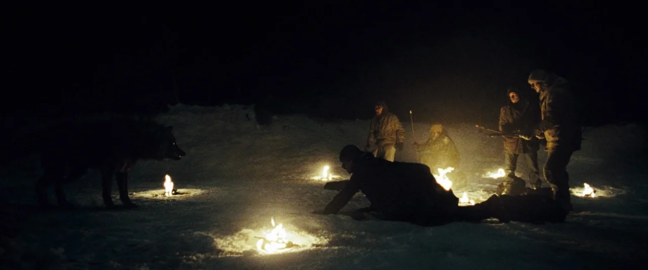The Grey (2012) movie still: wide — A group of men crouch around small fires burning directly on the snow at night. A large wolf stands at the edge…
