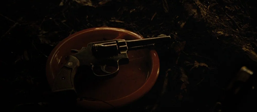The Road (2009) movie still: closeup — A dark, dirt-covered surface provides the backdrop for a plastic red plate. A metal revolver sits in the…