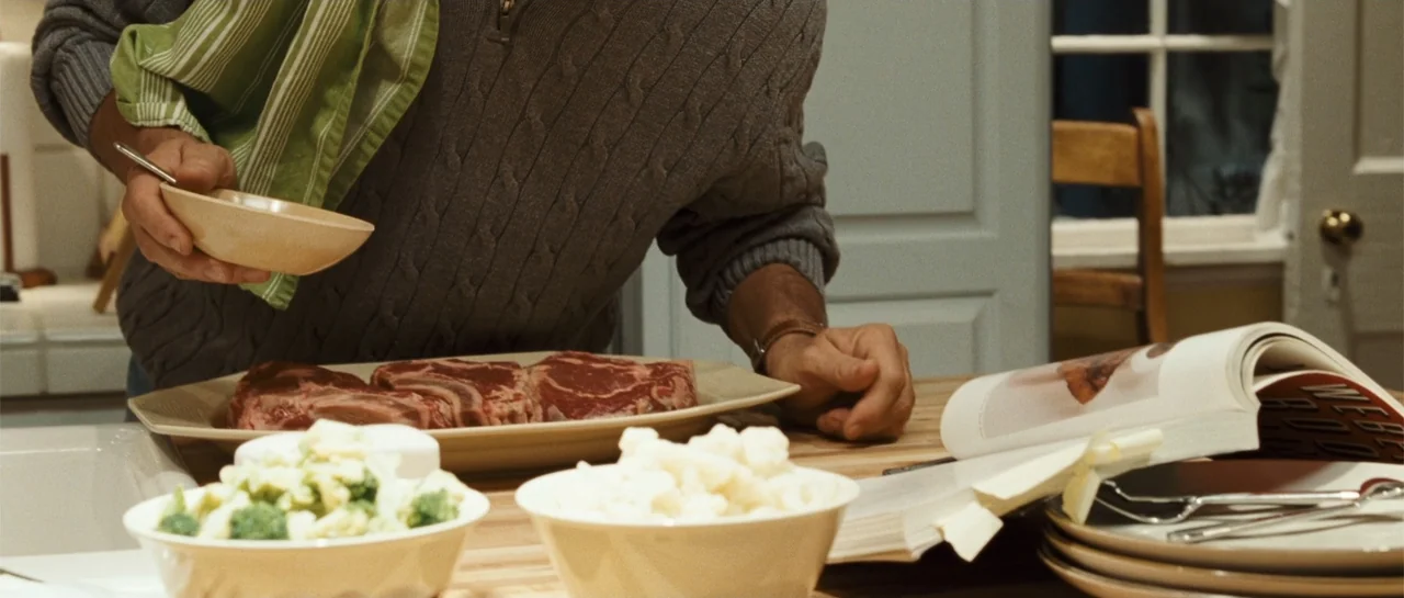 The New Daughter (2009) movie still: medium — A well-lit residential kitchen features a wooden table with raw steaks and prepared vegetables. A man…