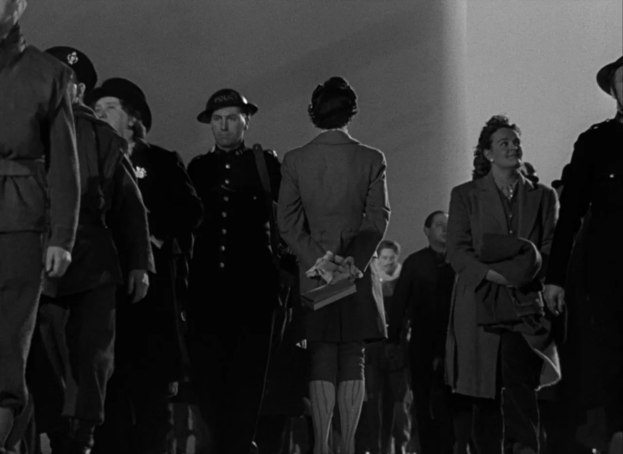 A Matter of Life and Death (1946) movie still: medium — A group of people walks together on a dark pavement at night under lighting. A woman…