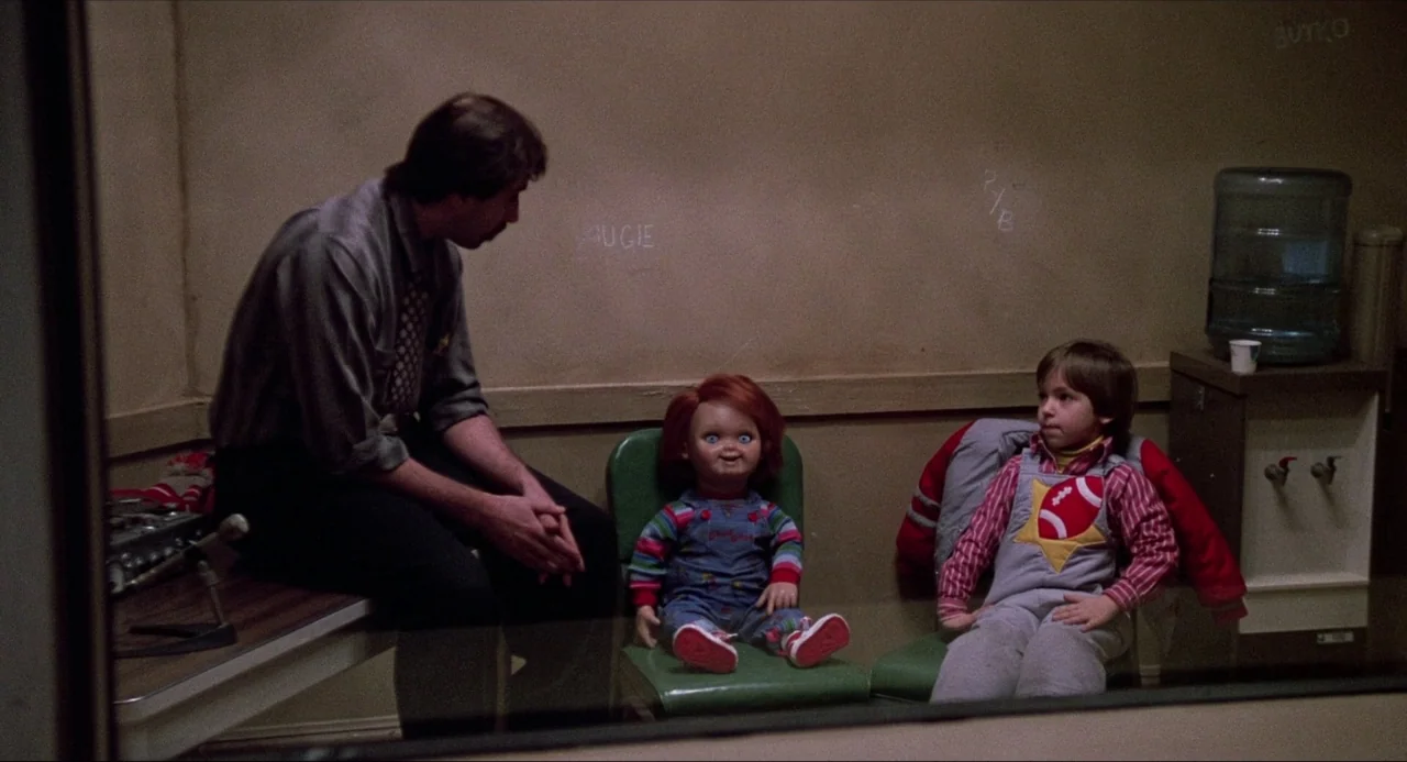 Child’s Play (1988) movie still: medium — An interrogation room with plain beige walls and a green plastic chair. An adult man sits next to a small…