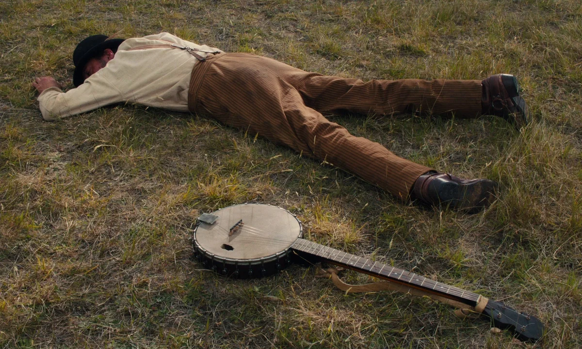 Slow West (2015) movie still: fullbody — A man lies prone in a dry, grassy field next to a discarded banjo. The open landscape shows sparse…