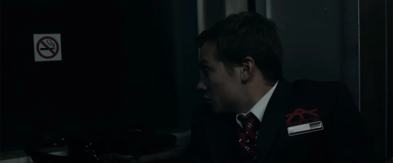 Howl (2015) movie still: medium — A dark, modern transportation interior features a wall with a prominent no-smoking sticker. A young man in a…