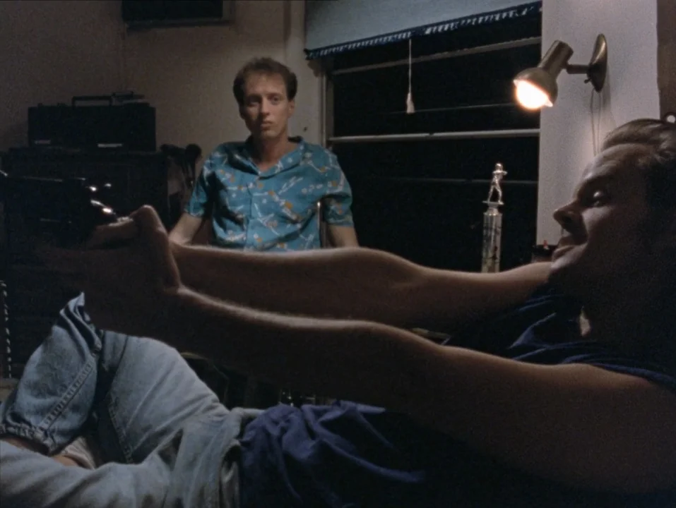 River of Grass (1994) movie still: medium — A dark, dimly lit room contains two young men at night. One man holds a handgun outstretched while…