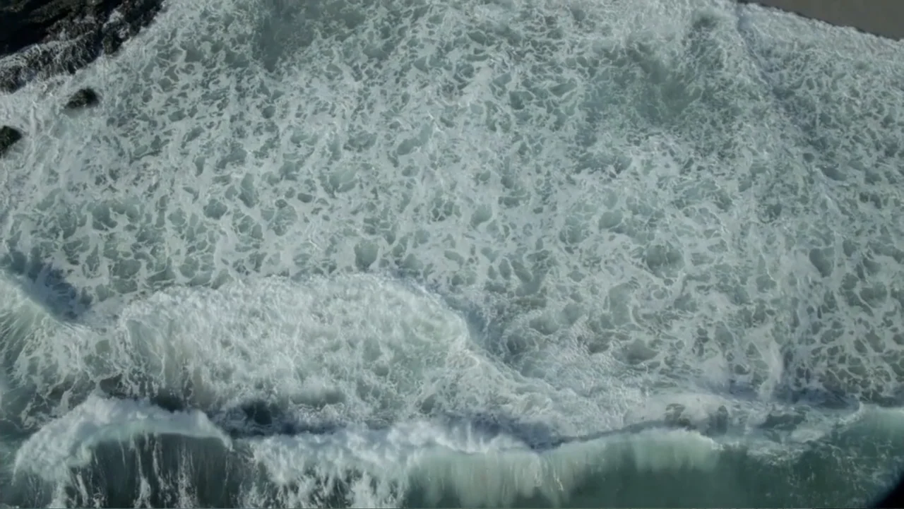 Empire Queen: The Golden Age of Magic (2024) movie still: establishing — An overhead view shows ocean waves breaking near a sandy beach. The water…