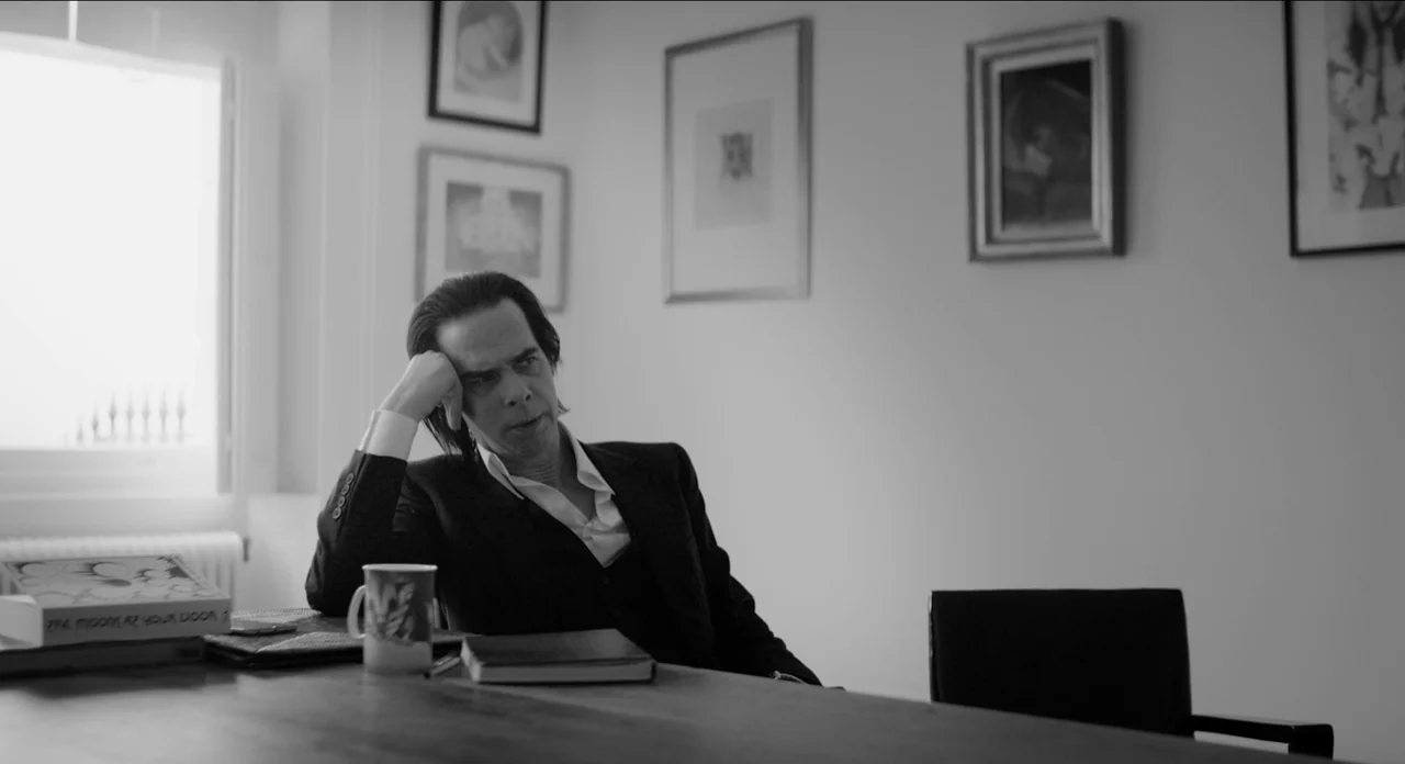 One More Time With Feeling movie still: medium — An office interior features framed artwork on a white wall and a wooden desk. A man in a suit sits…