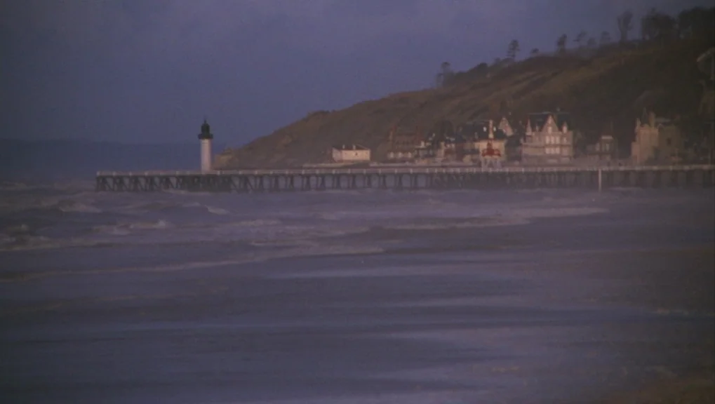Un Homme et Une Femme (1966) movie still: establishing — A long wooden pier extends into a dark, wavy sea under a purple twilight sky. A row of…