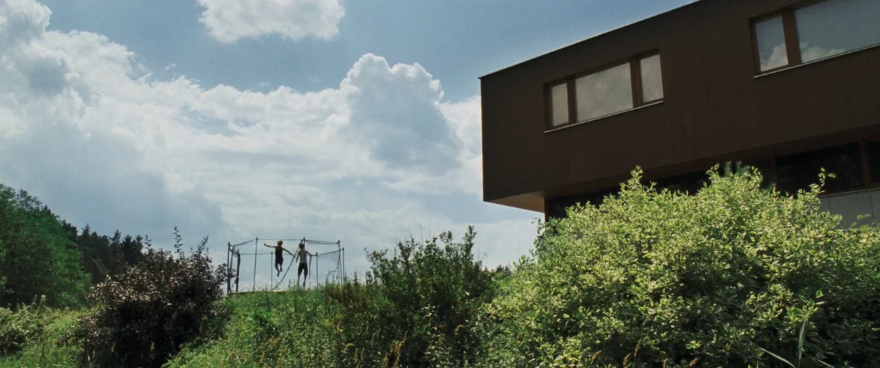 Goodnight Mommy (2014) movie still: wide — A modern two-story brown building stands beside a lush green field under white clouds. Two children jump…