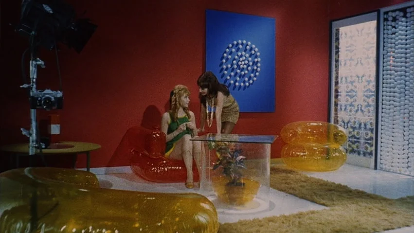 Four Times that Night (1971) movie still: medium — A 1960s interior with bright red walls and blue wall art with plastic furniture. Two women…