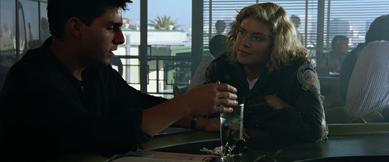 Top Gun (1986) movie still: medium — A sunlit diner interior with horizontal blinds obscuring an outdoor view of palm trees. A man gestures toward a…
