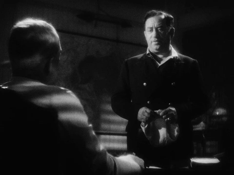 Island of Lost Souls (1932) movie still: medium — An interior space features dark walls and a map hanging in the background. A man stands wearing a…