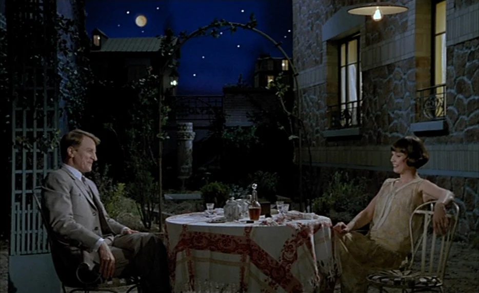 Mélo (1986) movie still: fullbody — A stone-walled courtyard features a small bistro table under a metal archway at night. A man and woman in 1920s…