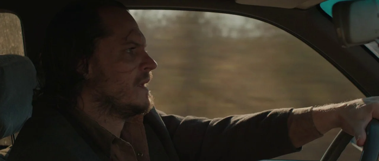 Joe (2013) movie still: medium — The interior of a moving vehicle shows the driver side from a side angle. A man with long hair and a beard drives…