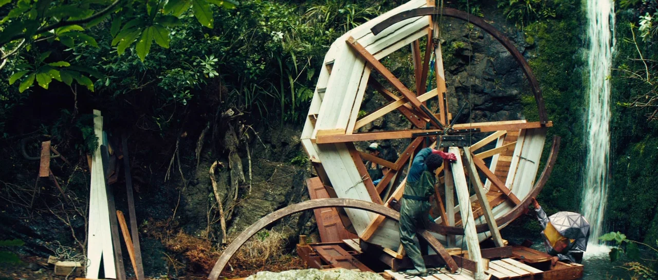 Z for Zachariah (2015) movie still: wide — A wooden water wheel frame sits at the base of a small rocky waterfall. Two workers in protective gear…