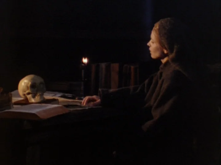 The Magic Flute (1975) movie still: medium — A dark, wood-paneled room features a heavy desk with open books and a candle. A woman sits in profile,…