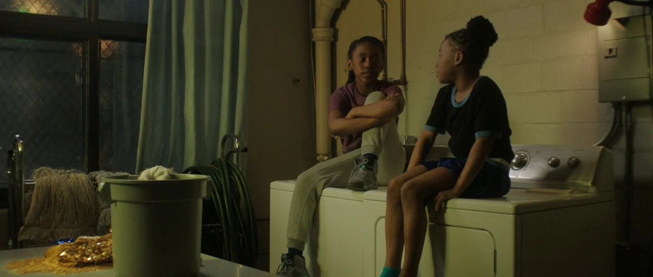 The Fits (2016) movie still: medium — A basement laundry room features painted concrete walls, exposed piping, and white appliances. Two young girls…