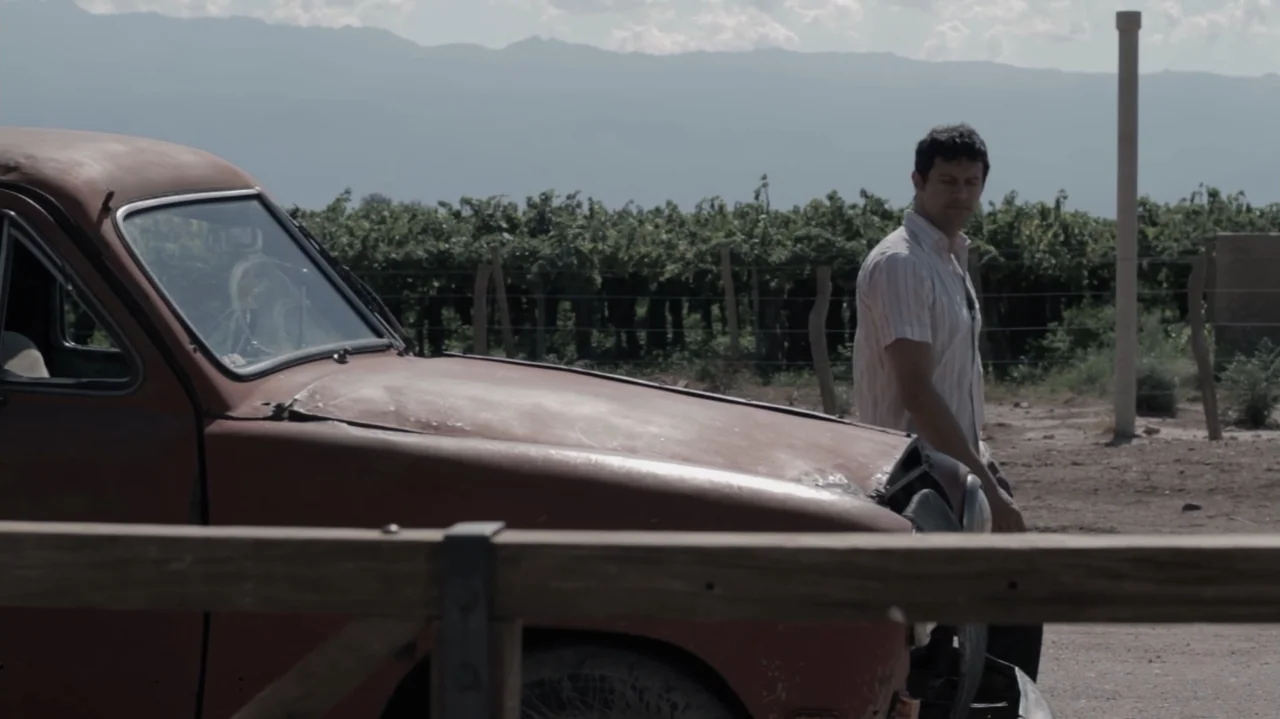 Still Life (2013) movie still: medium — A rural vineyard landscape features mountains in the distance and rows of grape vines. A man in a striped…