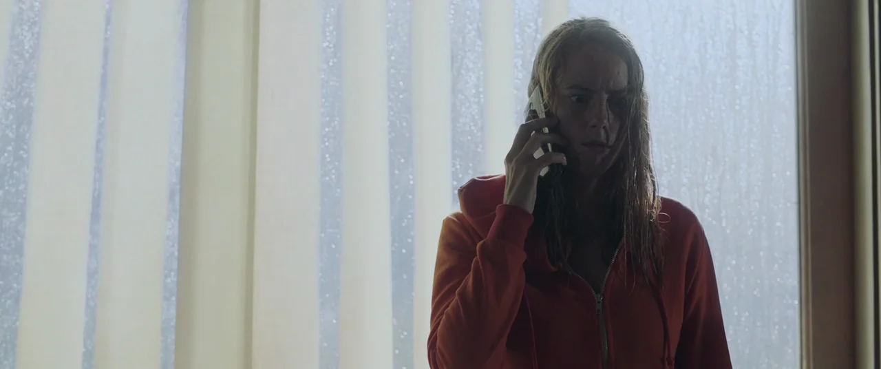 Crawl (2019) movie still: medium — A woman stands before a window covered by pale vertical blinds during rain. She holds a smartphone to her ear…