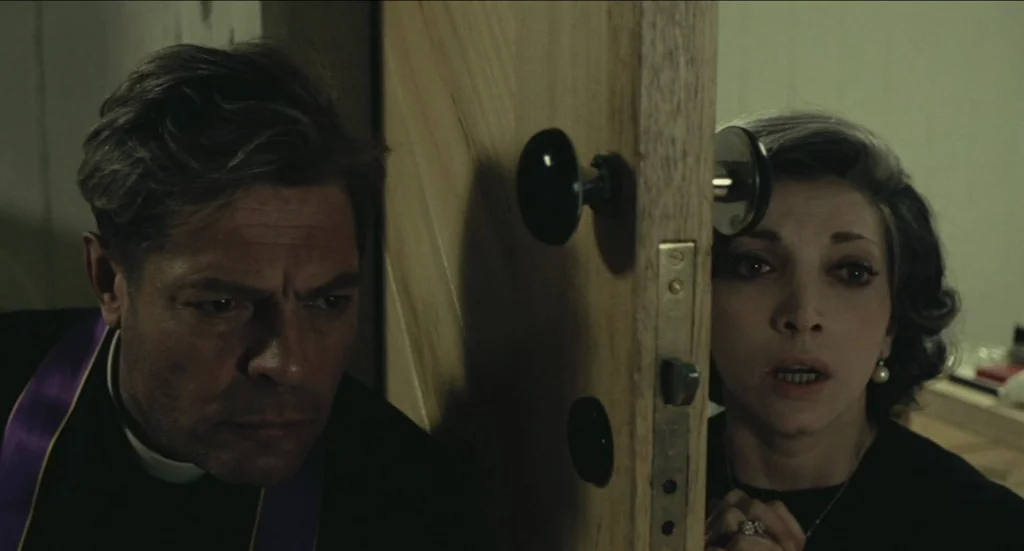 Todo Modo (1976) movie still: medium — A dark, narrow interior space is bisected by a wooden door held slightly open. A priest and a woman peer…