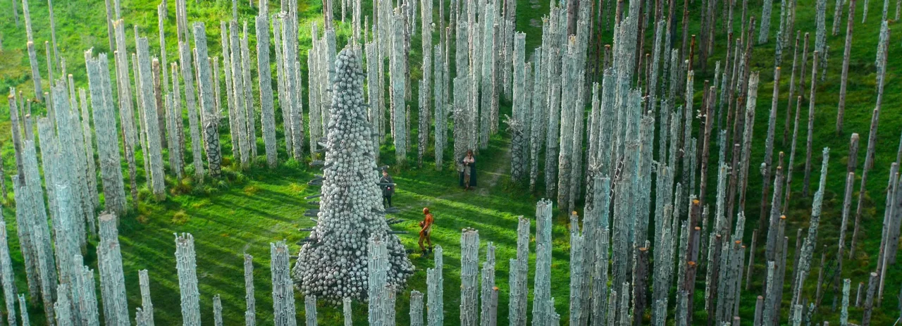 28 Years Later (2025) movie still: wide — A clearing in a grassy field contains numerous pale vertical wooden poles arranged systematically. A tall,…