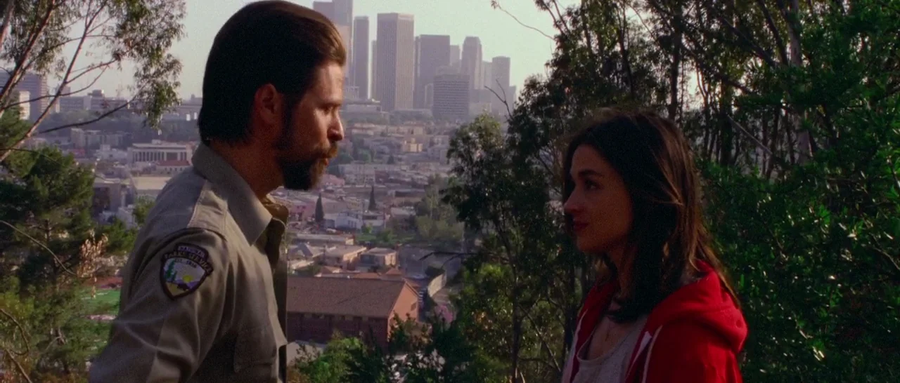 Too Late (2015) movie still: medium — A grassy hillside overlooks a sprawling cityscape with multiple skyscrapers in the distant background. A…