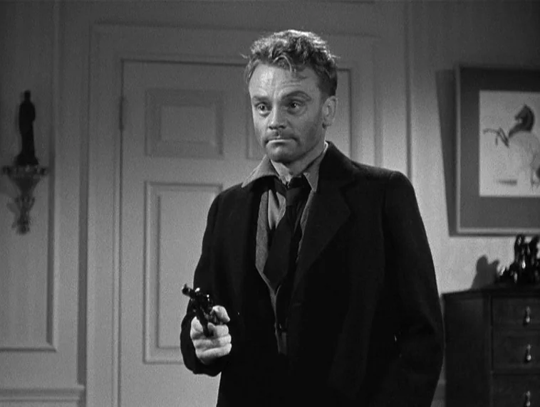 The Roaring Twenties (1939) movie still: medium — A starkly furnished room features a paneled door and a framed wall hanging. A man stands in the…