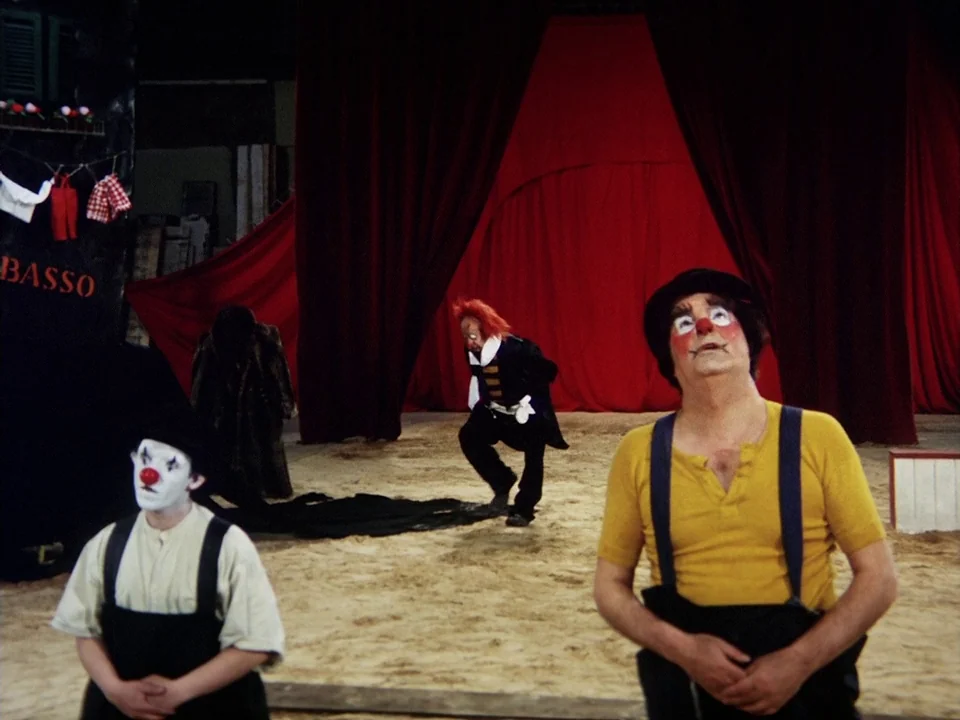 The Clowns (1970) movie still: fullbody — An indoor circus ring with sand on the floor and large deep red curtains. Three adult male clowns wearing…