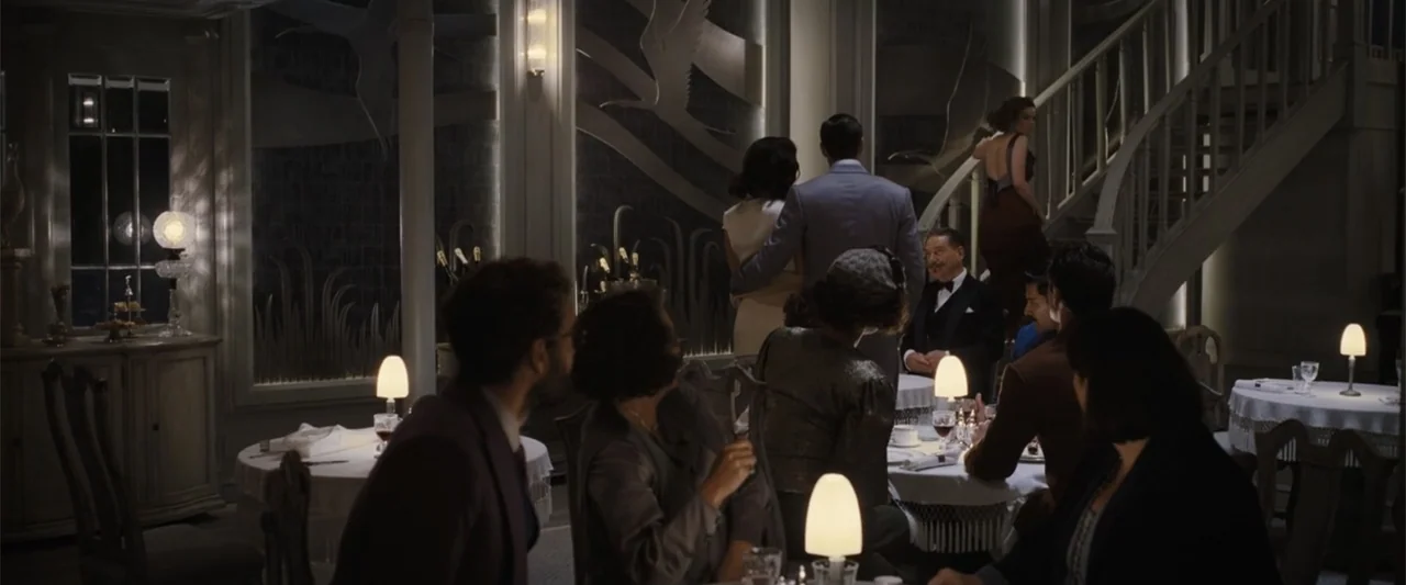Death on the Nile (2022) movie still: wide — A 1930s style dining room with an elegant staircase and decorative wall panels. Several people sit at…