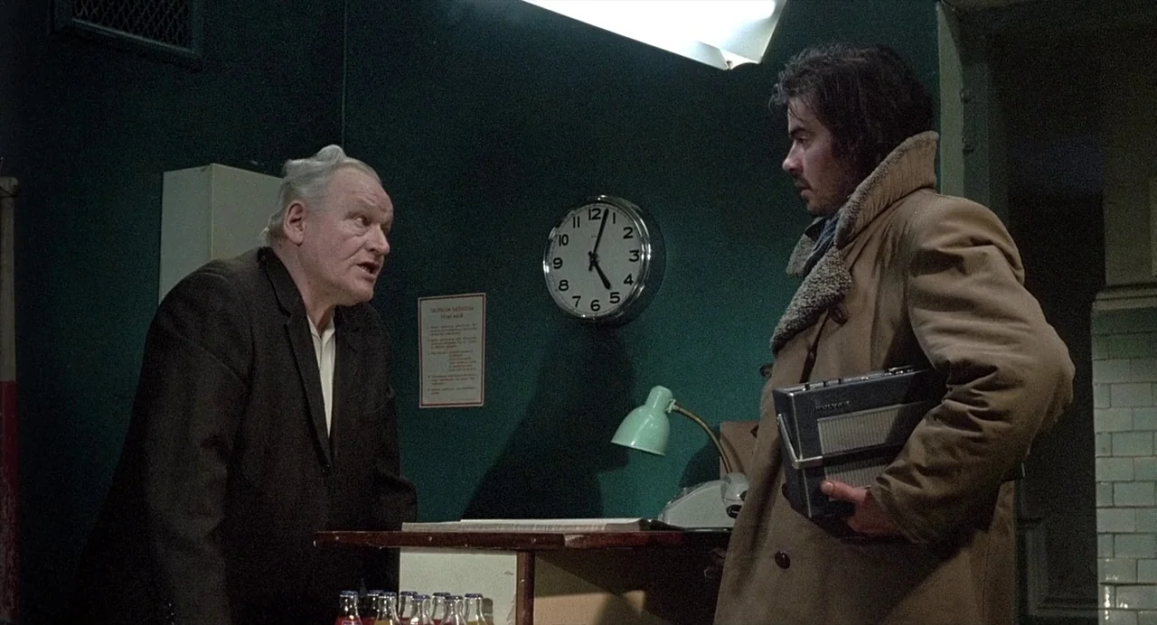 Ariel (1988) movie still: medium — A dimly lit room features dark green walls, a wall clock, and a desk. An older man talks to a younger man who…