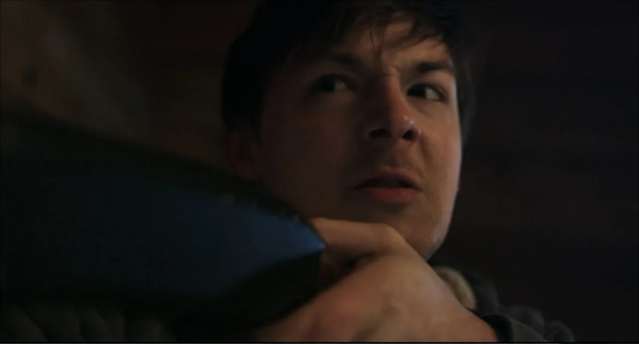 Freedom (2024) movie still: closeup — A dark indoor space with minimal light and obscured architectural details. A young man stares intently while…