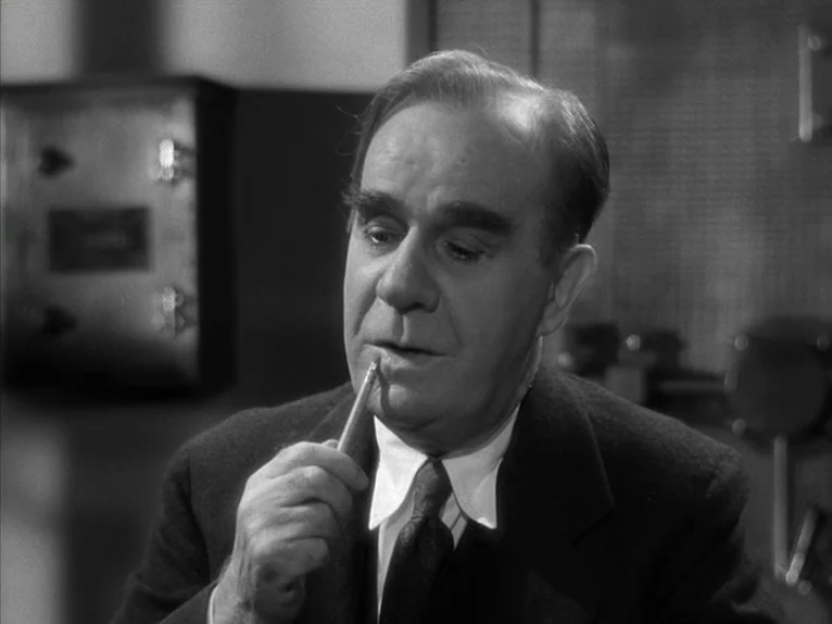 Shadow of a Doubt (1943) movie still: closeup — A man sits in a dimly lit space with metallic industrial equipment in background. He looks down…