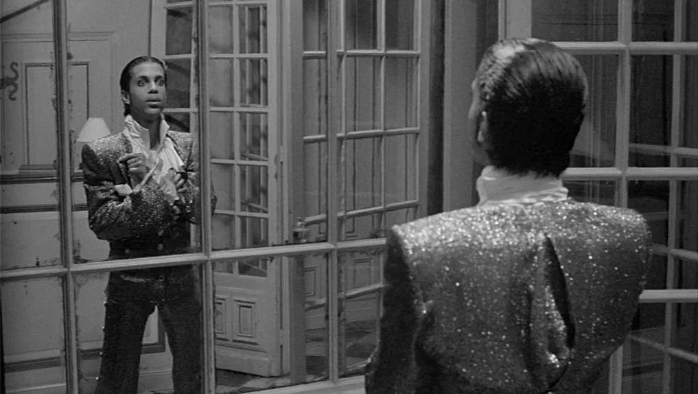 Under The Cherry Moon (1986) movie still: medium — A man stands before a large paneled mirror inside a well-lit dressing area. He gazes directly…