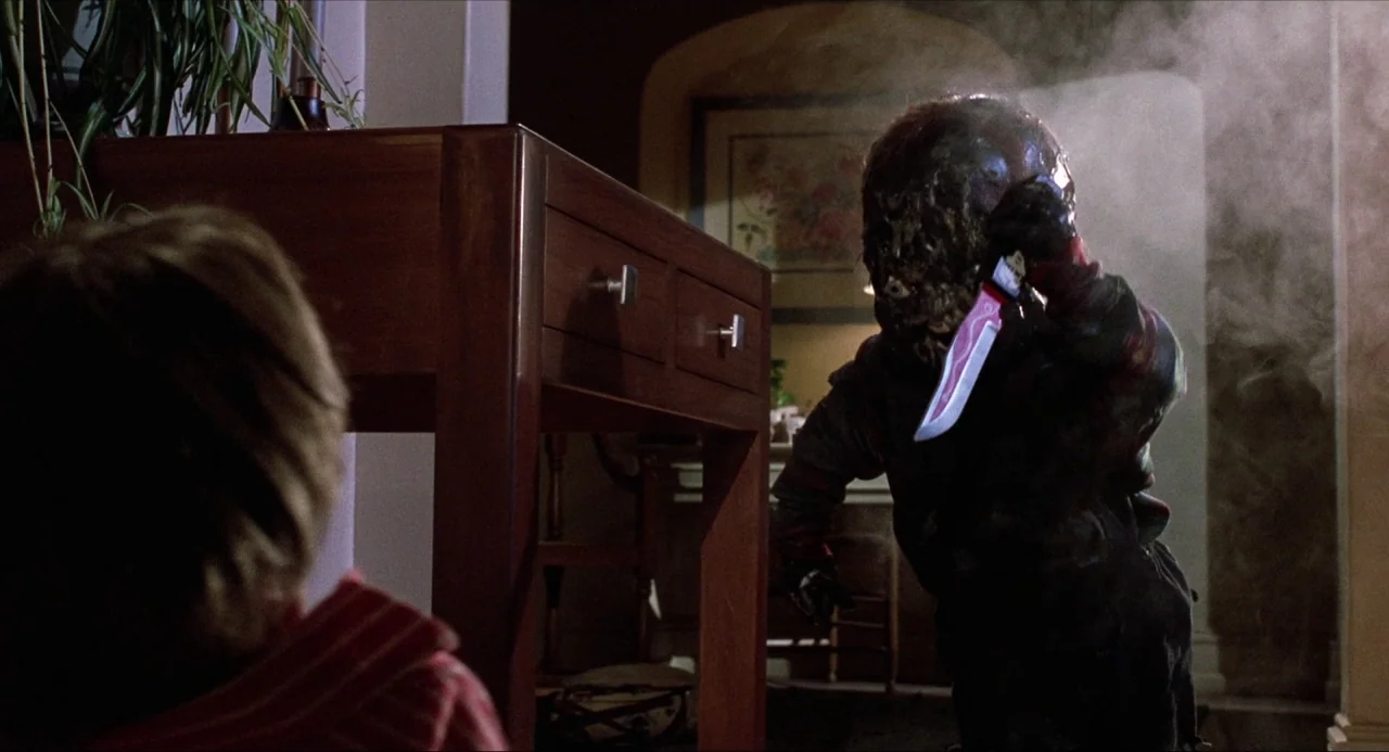 Child’s Play (1988) movie still: medium — A dimly lit indoor hallway with a wooden table in the immediate foreground. A small, burned figure…
