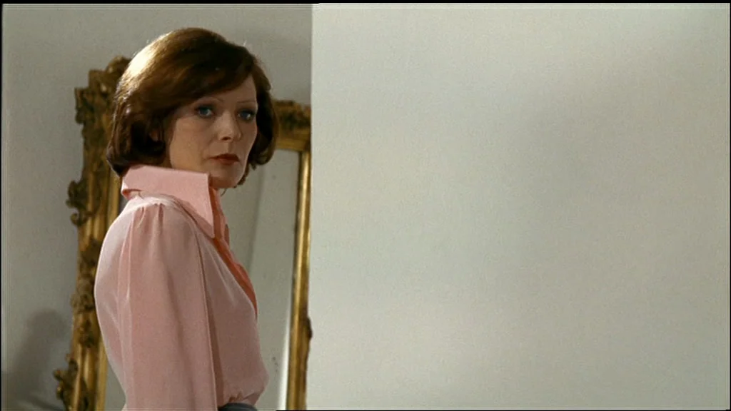 Chinese Roulette (1976) movie still: medium — An interior space features a plain white wall and a decorative gold-framed mirror. A woman in a pink…