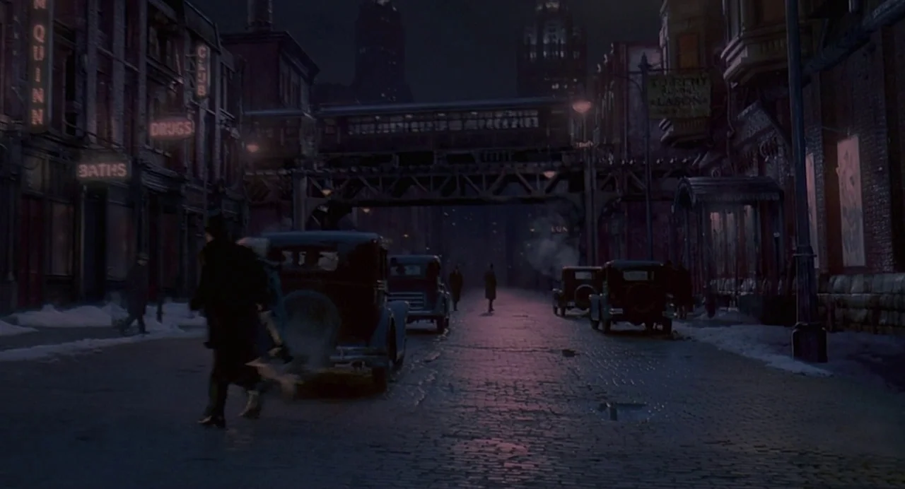 Chicago (2002) movie still: establishing — A dark, snow-covered cobblestone city street framed by old brick buildings and neon signs. An elevated…