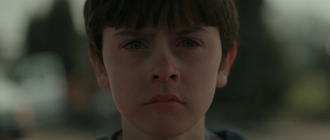 Bullhead (2011) movie still: closeup — A young child stands outdoors under overcast daylight conditions. The child looks directly into the camera…