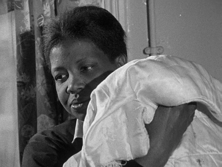Cathy Come Home (1969) movie still: closeup — A dimly lit indoor space with a patterned curtain in the background. A young woman holds a folded…