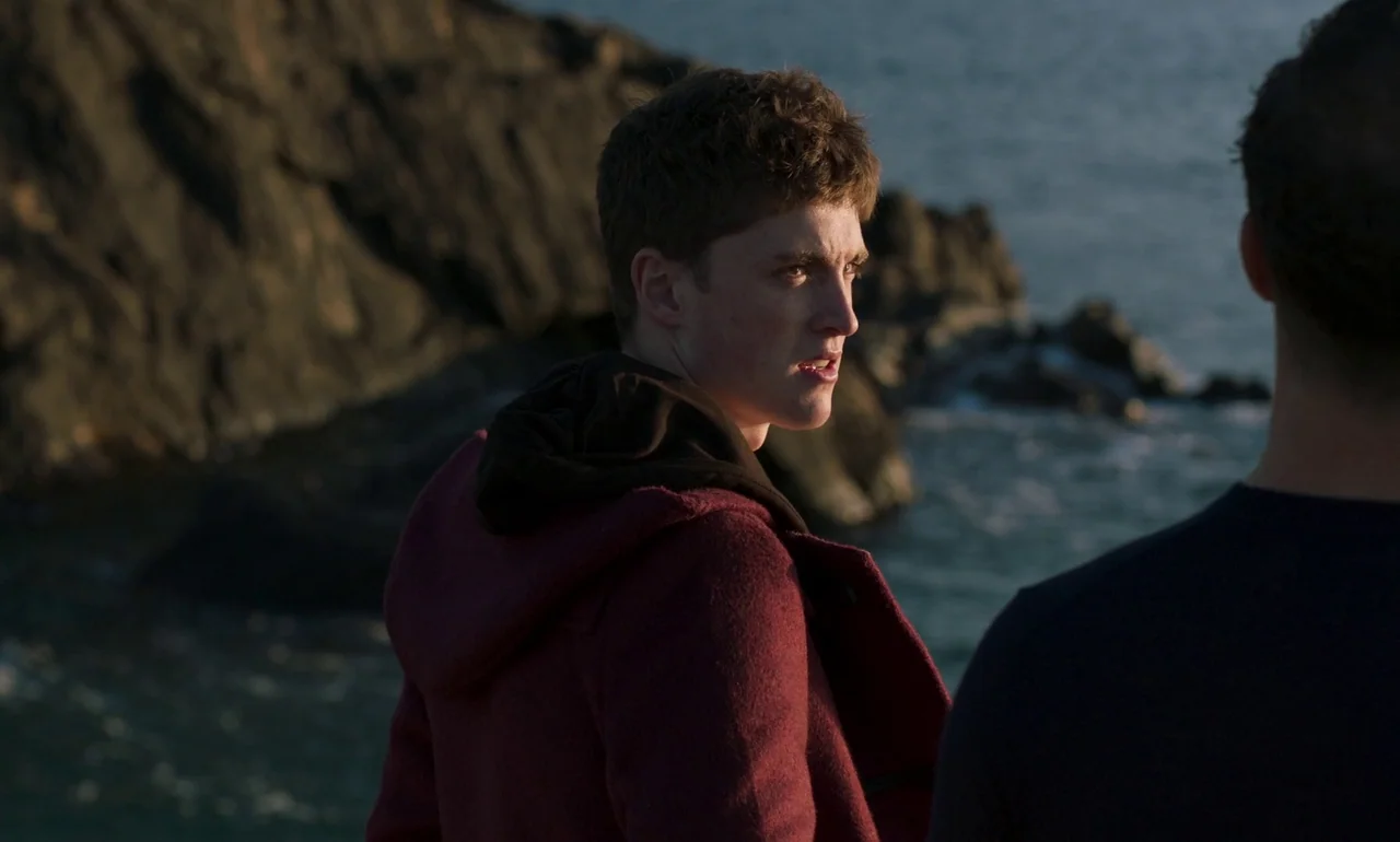 It Is In Us All (2022) movie still: medium — Two men stand near a dark rocky cliffside overlooking the ocean at sunset. One man in a burgundy coat…