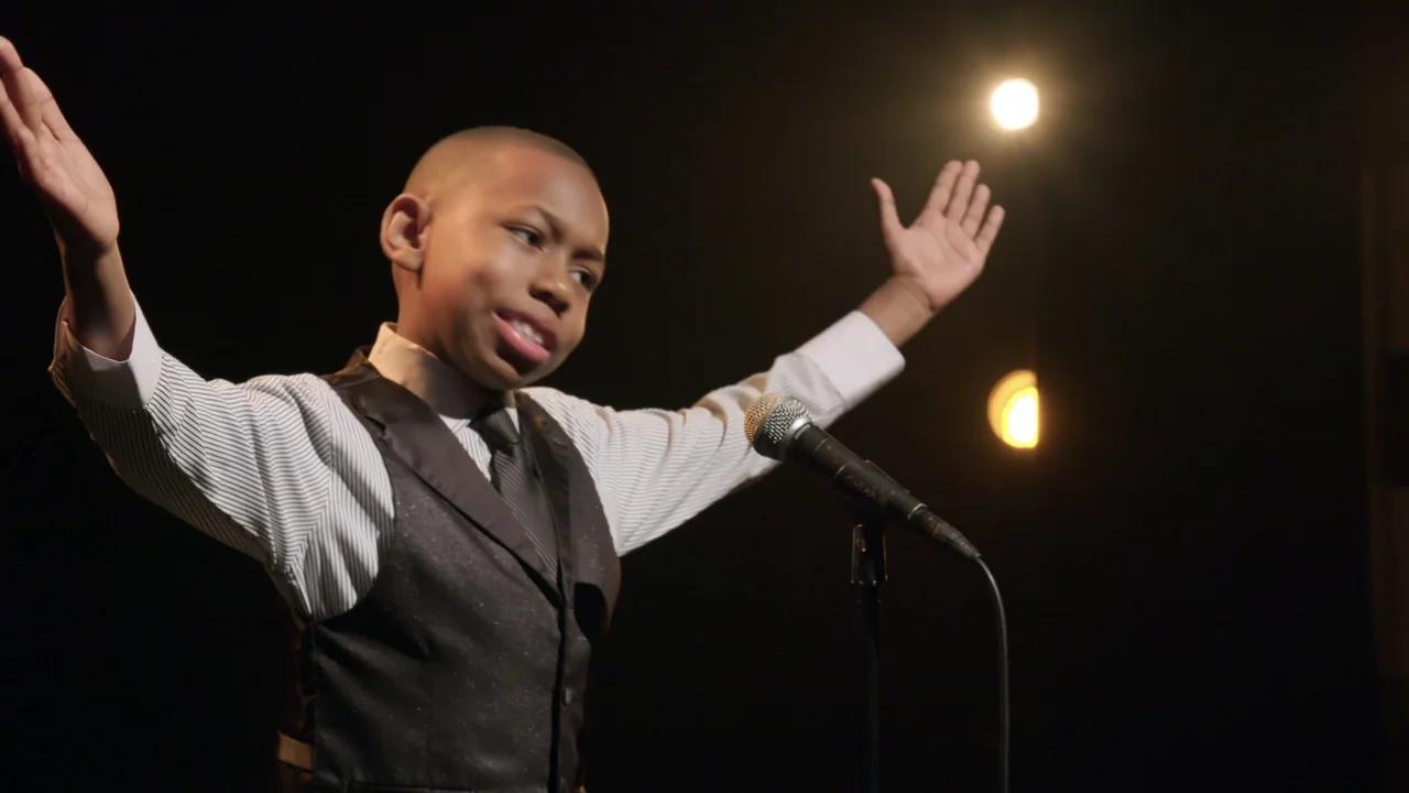 We Are the Dream: The Kids of the Oakland MLK Oratorical Fest (2020) movie still: medium — A dimly lit stage features a lone boy in formal attire…