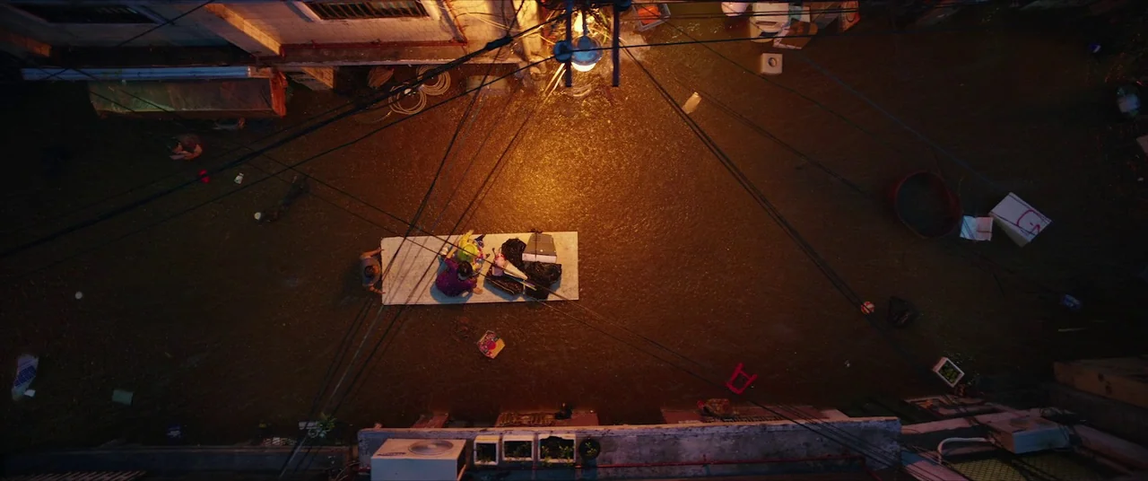 Parasite (2019) movie still: wide — A dark flooded alleyway at night holds debris and thick, murky brown water. Two people sit on a floating…