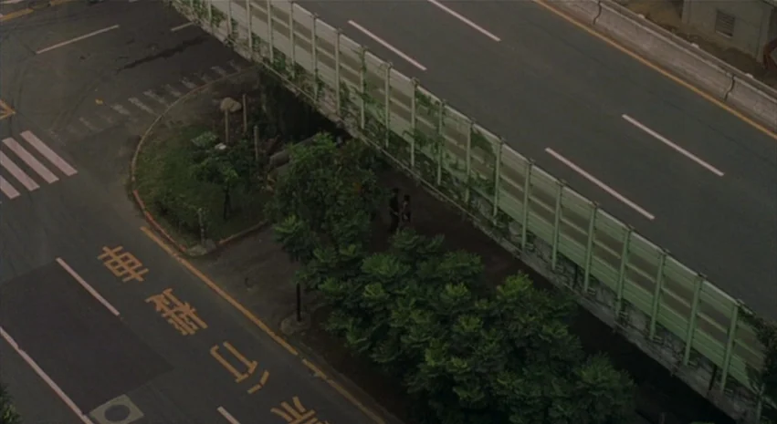 Yi-Yi (A One and A Two) (2000) movie still: wide — A high-angle view of a multi-lane roadway adjacent to a concrete sound barrier wall. Two figures…